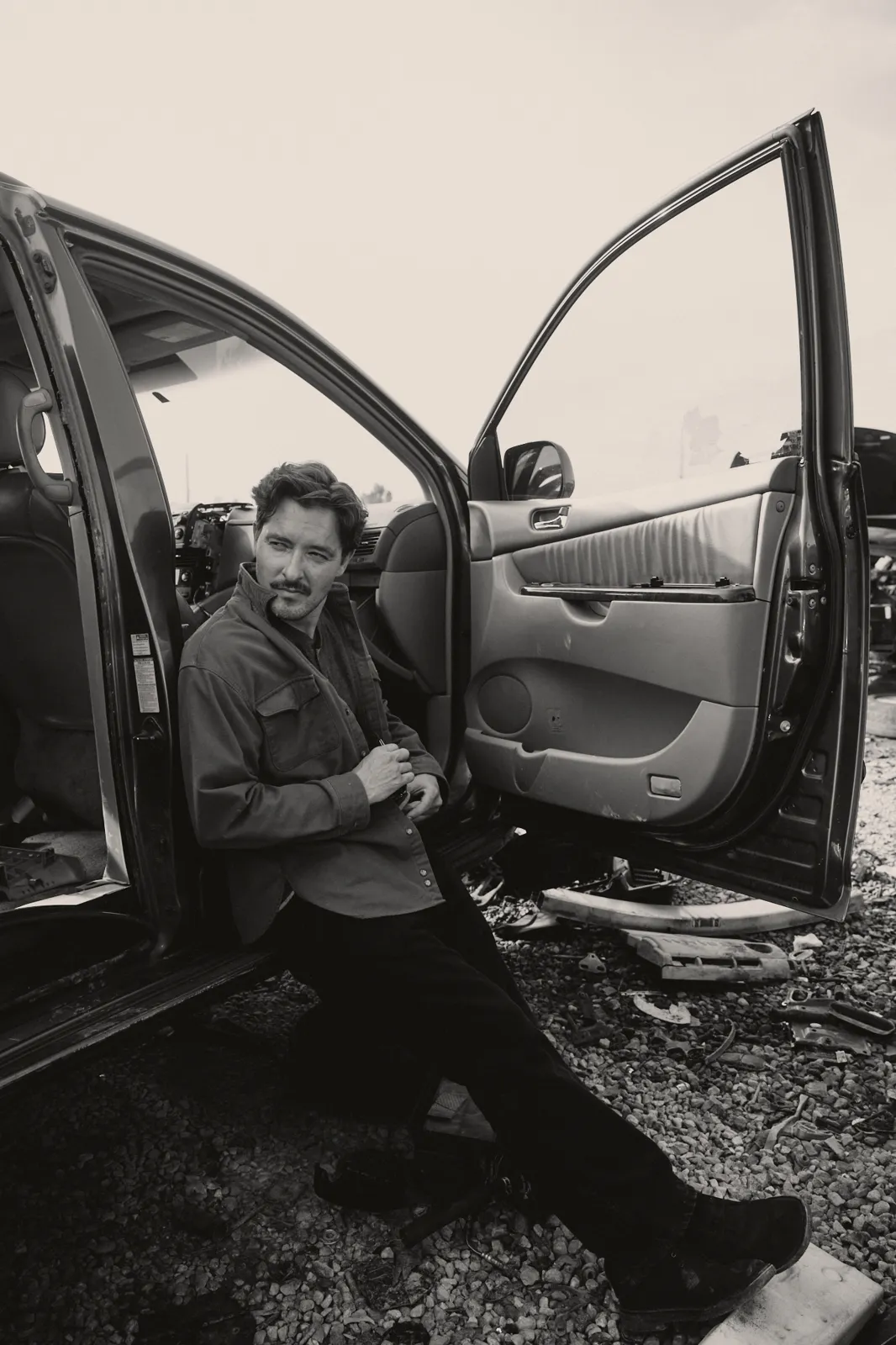 Black-and-white photo of Graham Hill sitting in an open car door at a junkyard