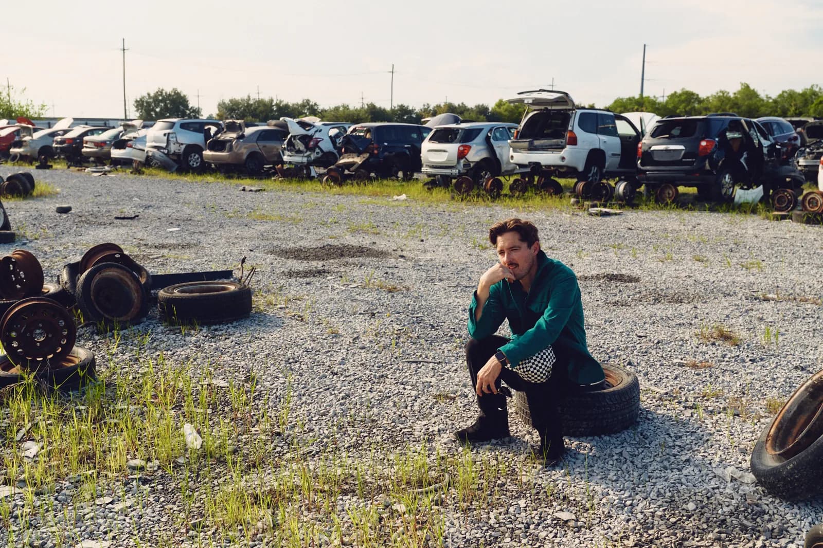 Graham Hill crouched on a tire in a gravel junkyard lined with cars